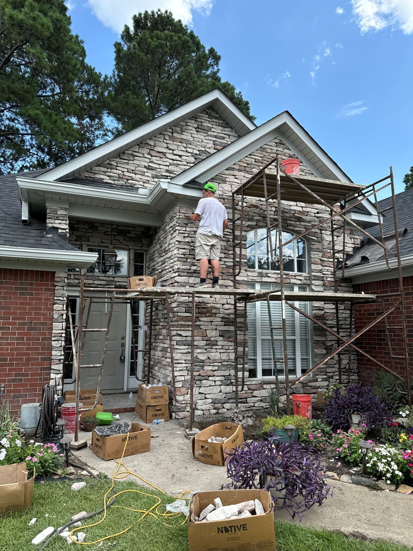 Worker on scaffolding repairing a brick house facade with stone trim and flower beds below