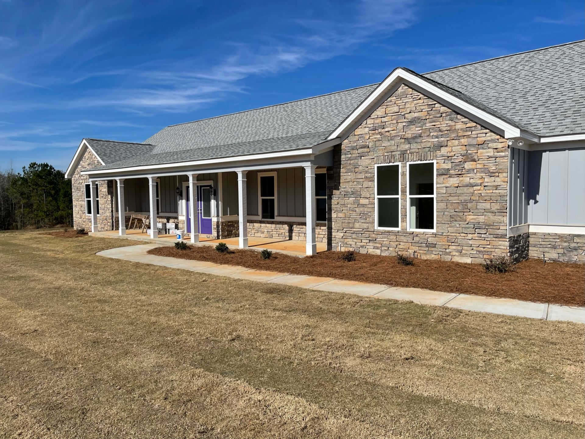 Single-story stone house with a white porch, gray roof, and brown lawn under a blue sky