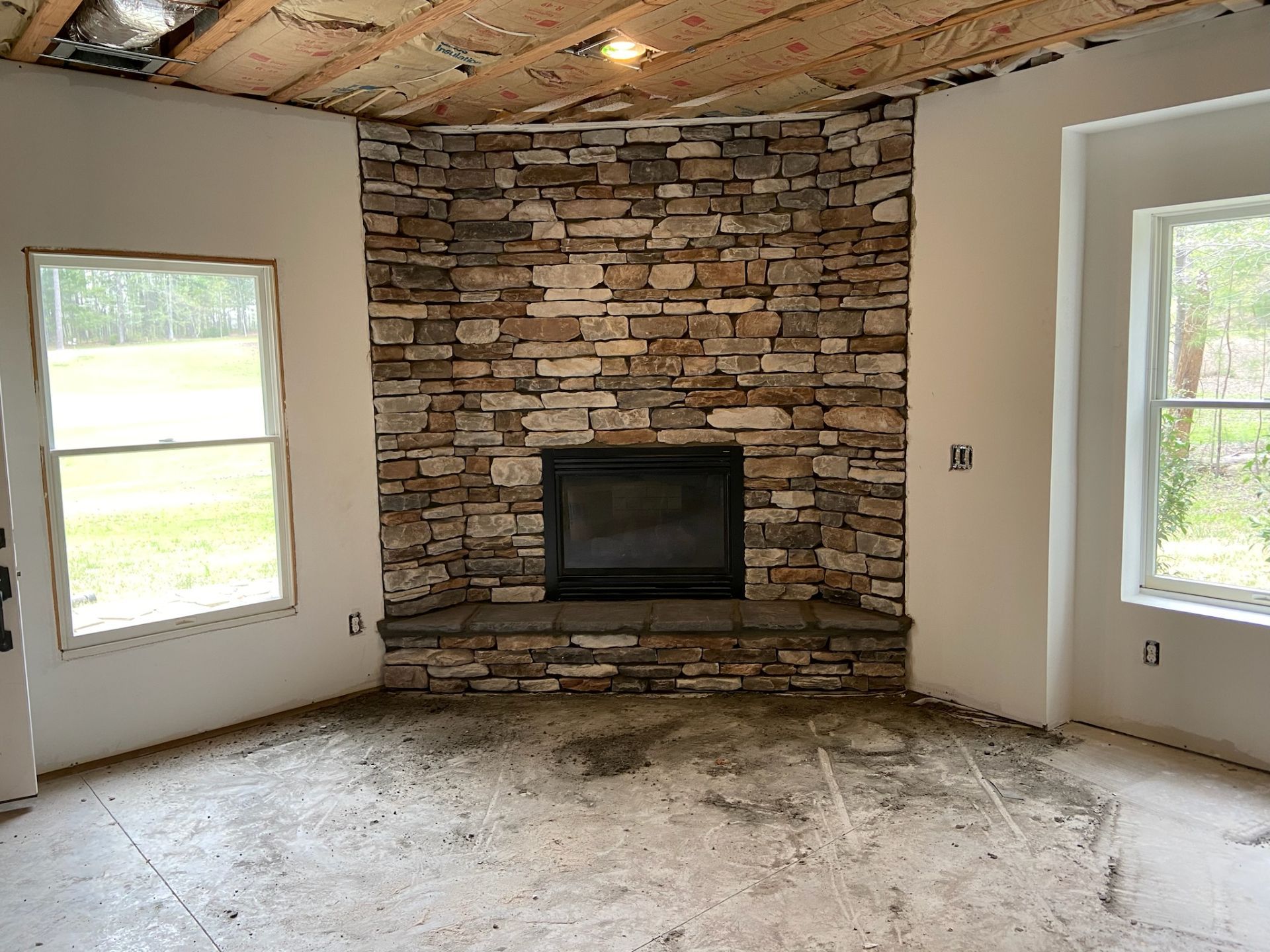 Living room with stone fireplace, two windows, unfinished ceiling, and bare floor