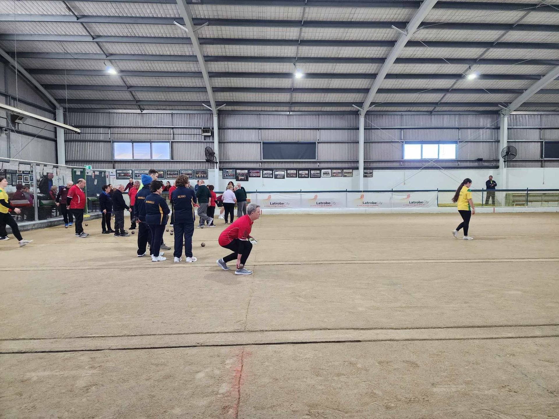 People playing indoor bocce on a dirt court in a large sports hall, with spectators nearby