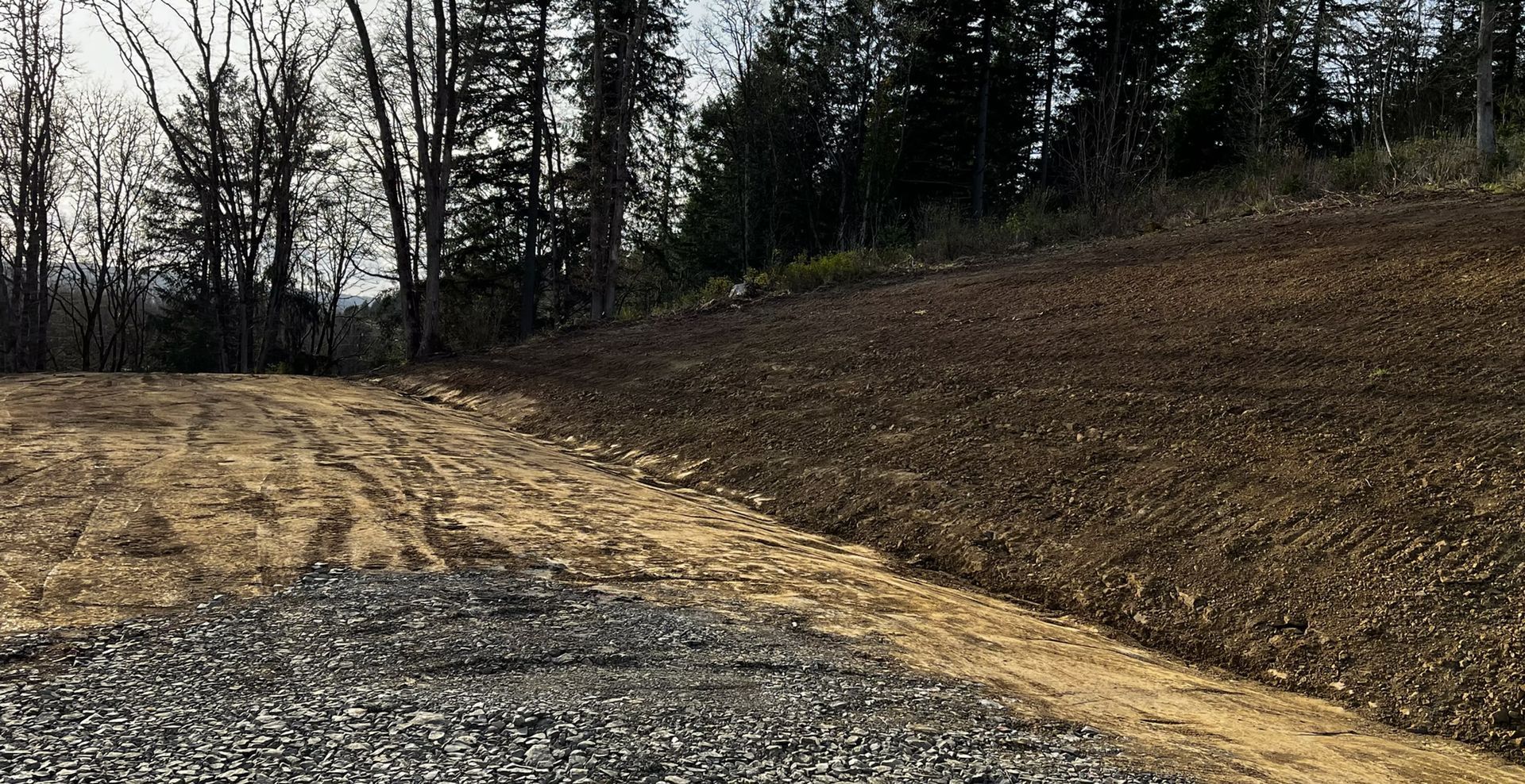 Cleared gravel and dirt lot beside a wooded hillside under overcast sky