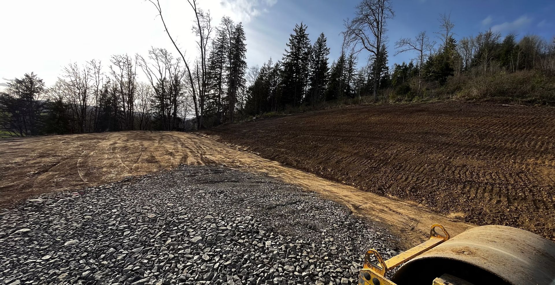 Gravel lot beside a dirt berm under clear sky, with bare trees and a yellow machine edge in the foreground