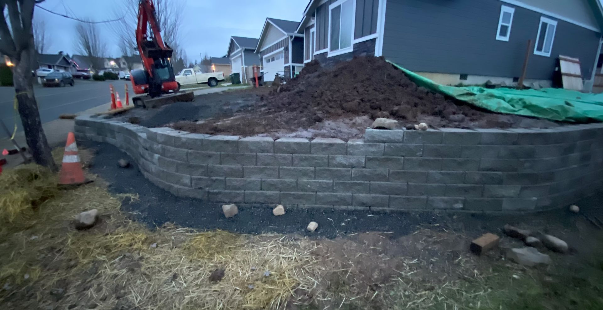 Residential construction site with a retaining wall, dirt mound, and excavator beside a house.