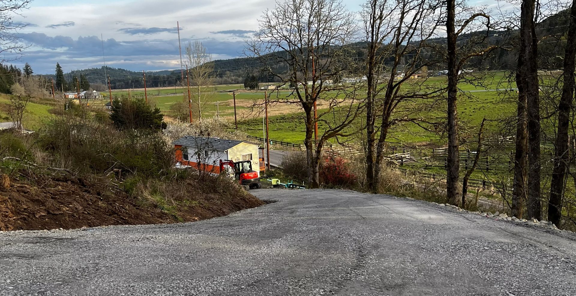 Gravel road curving downhill past a small house and fields under a cloudy sky