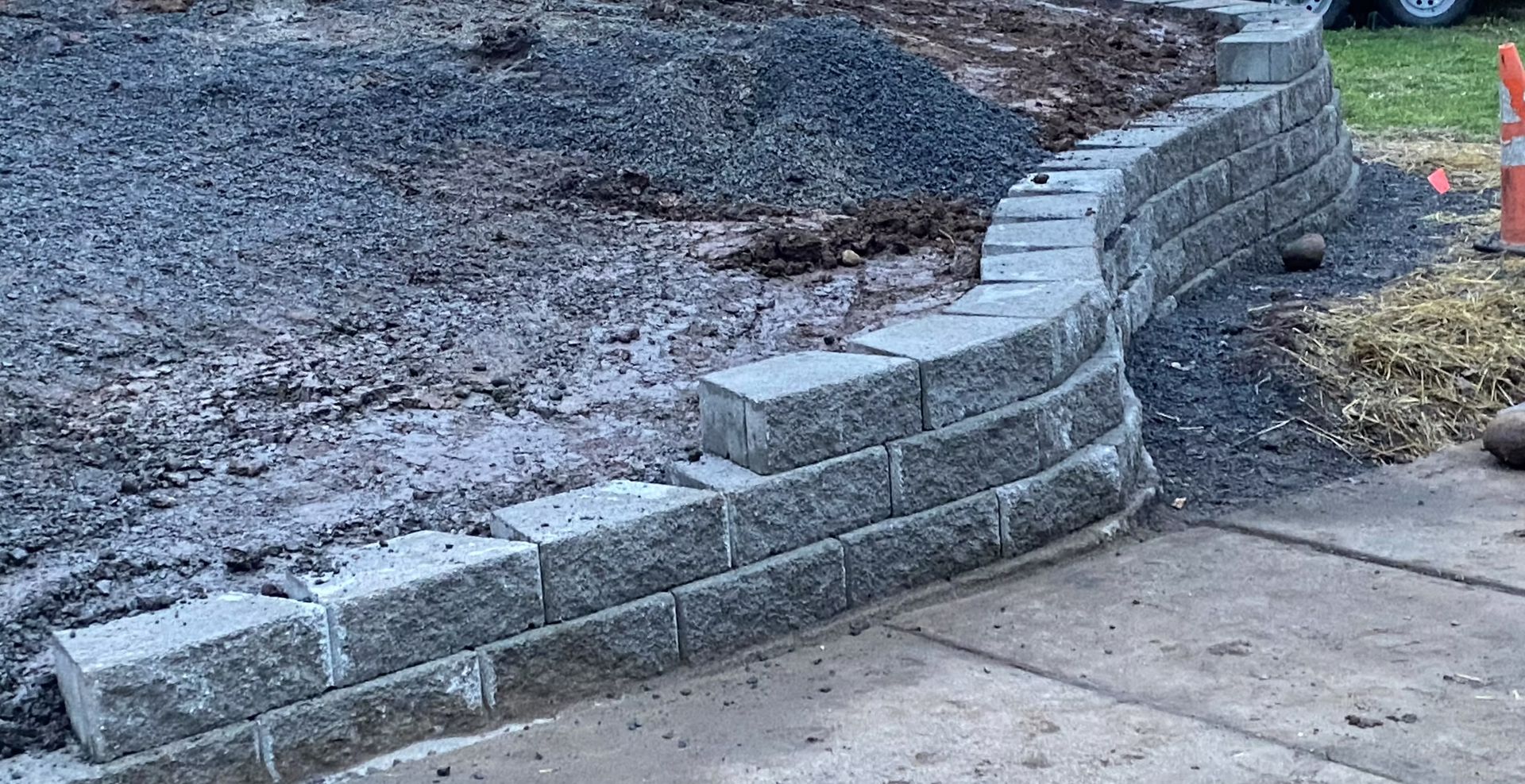 Curved gray block retaining wall beside a sidewalk and dirt lot, with orange cones in the background