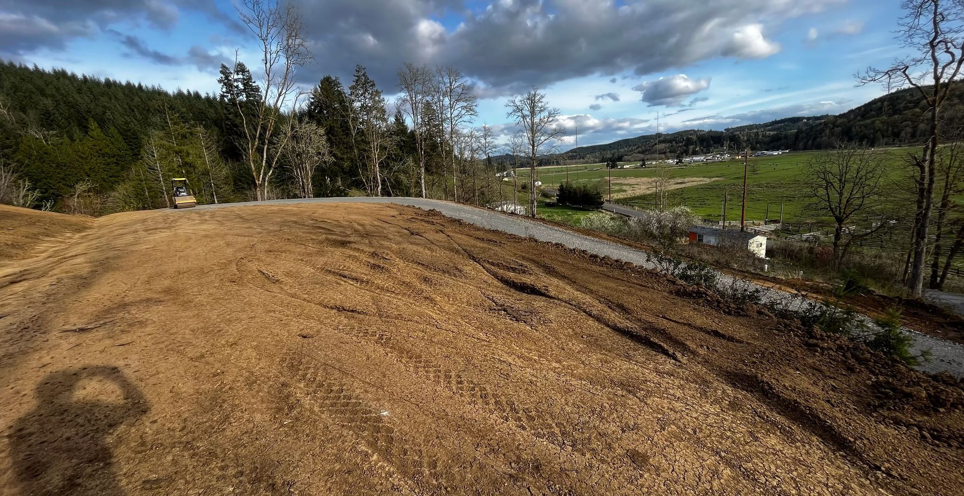 Freshly graded dirt mound beside a rural path, with trees, fields, and cloudy sky in the background