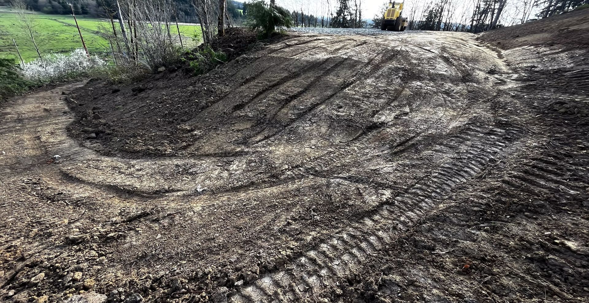 Mud-covered slope with tire tracks and a small excavator in the background under a cloudy sky