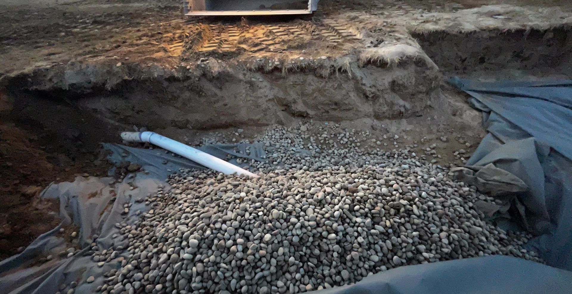 Excavated trench filled with gravel, black sheeting, and a white pipe on the left