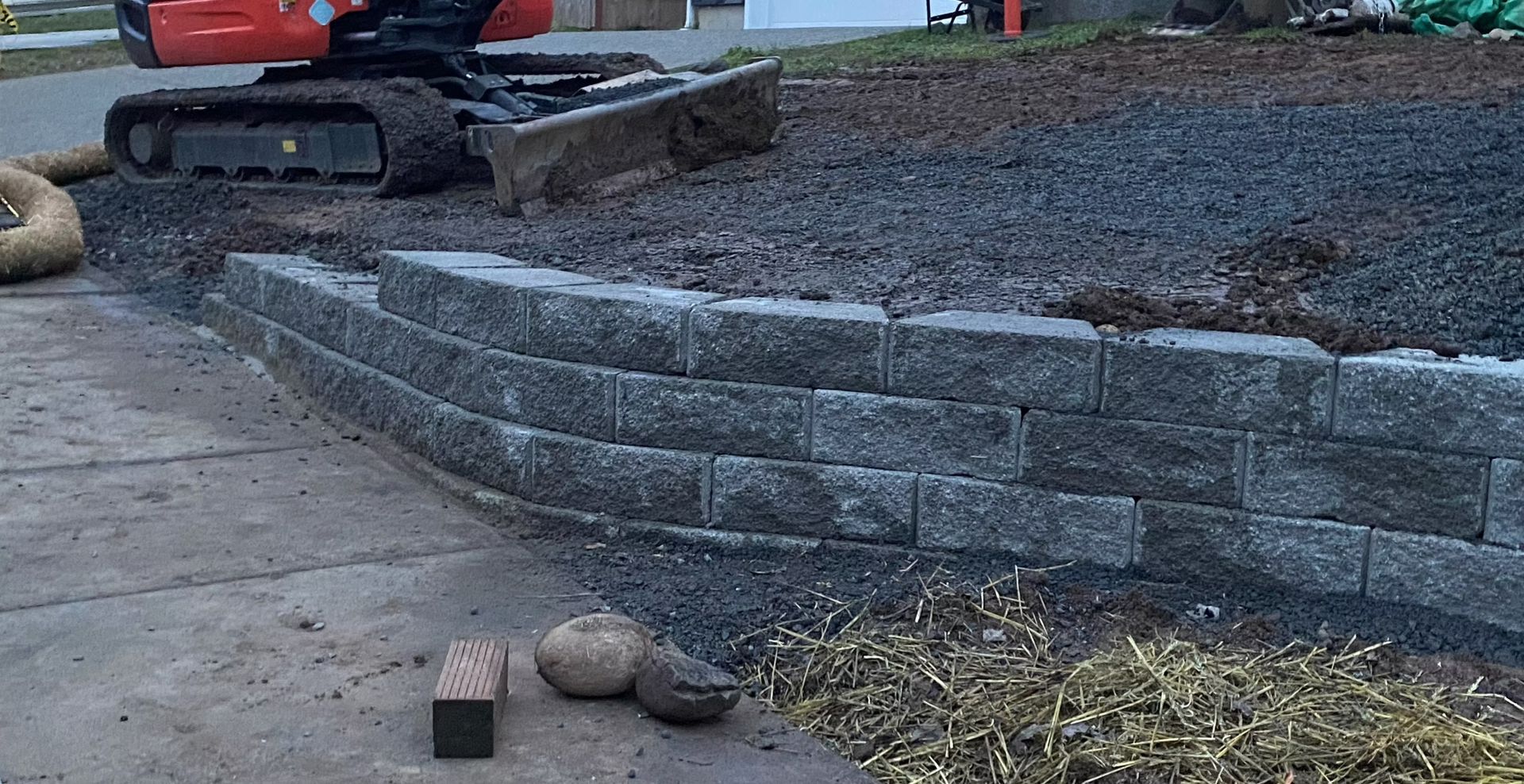 Construction area with a small excavator beside a curved gray retaining wall and dirt slope