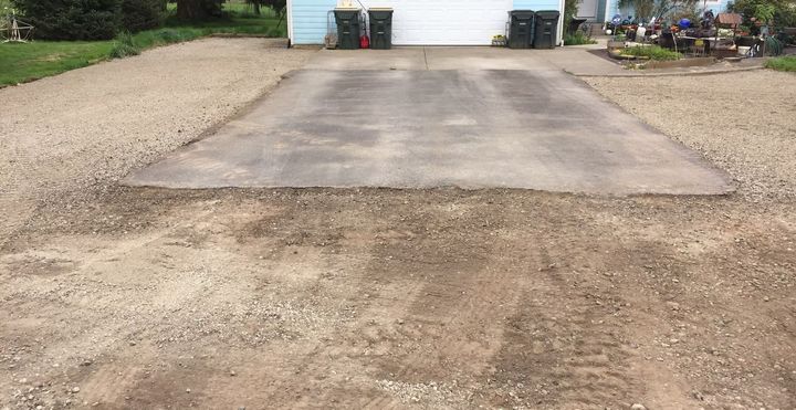 Concrete driveway leading to a garage, bordered by gravel and dirt in a rural yard.