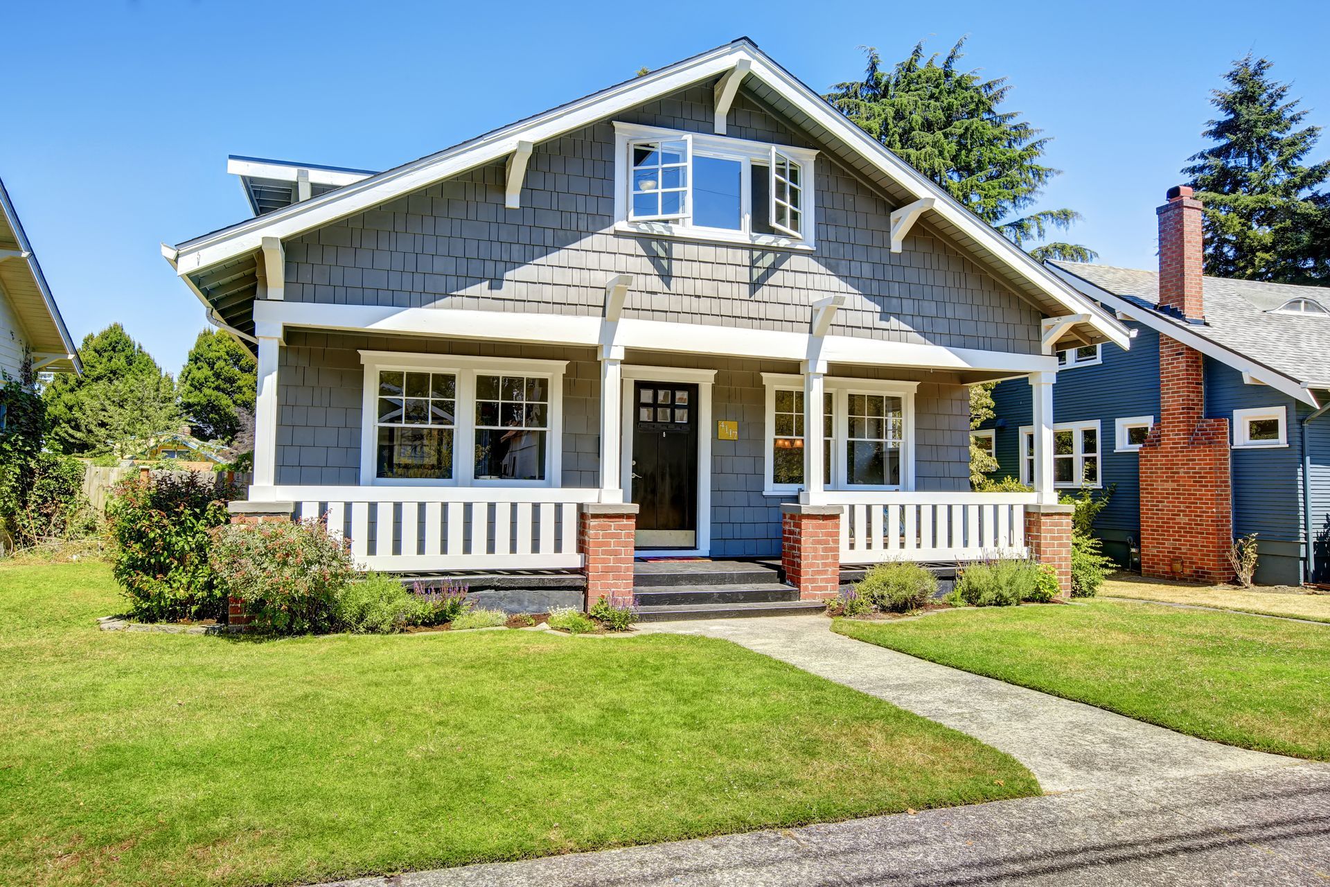 Gray Craftsman-style house with white trim, porch, and a brick chimney. Green lawn and blue sky.