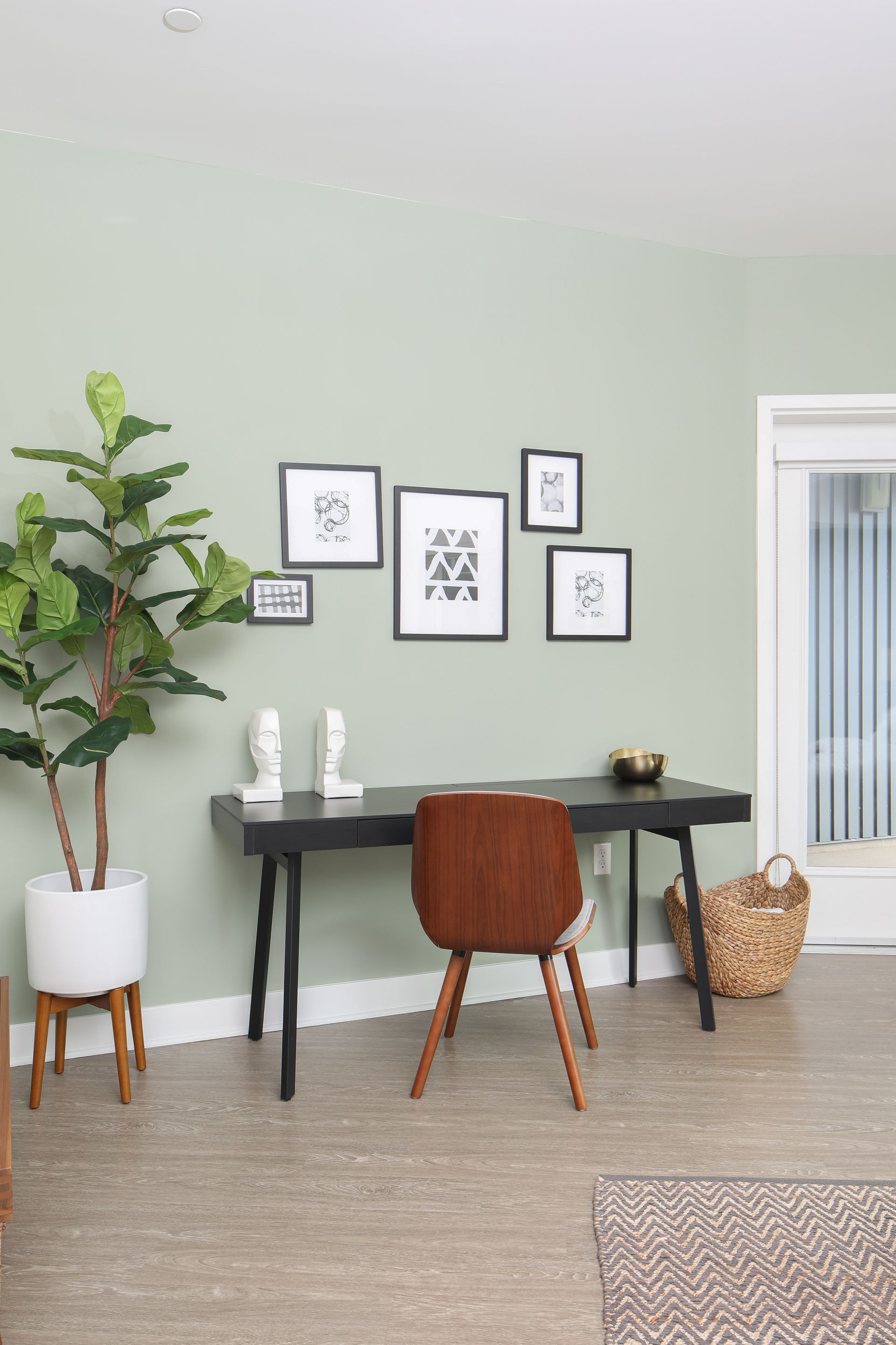 Minimal home office with black desk, wooden chair, wall art, plant, and woven basket against pale green walls