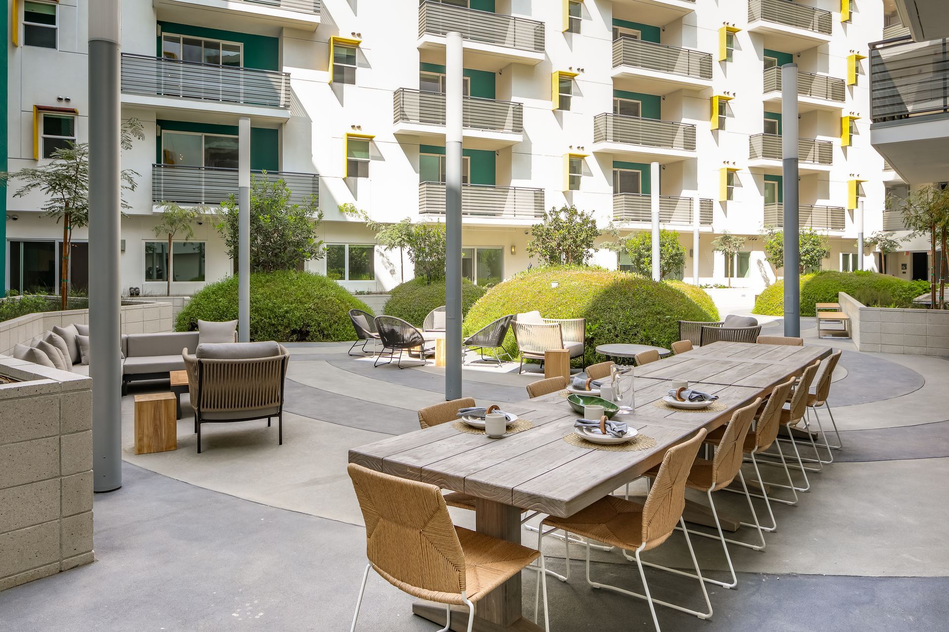 Outdoor courtyard with long wooden dining tables and chairs beside modern apartment buildings and greenery