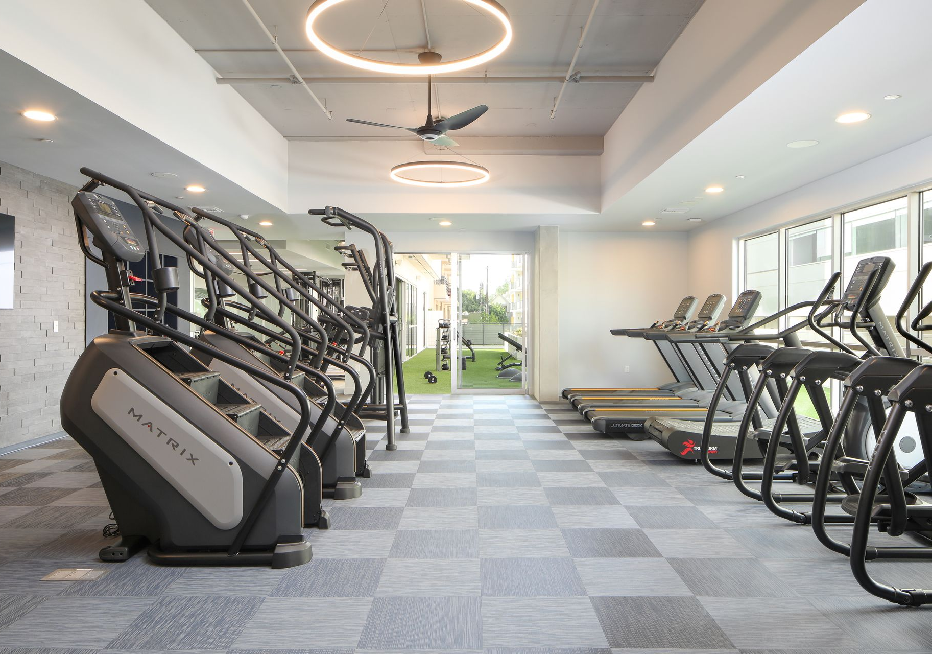 Modern gym with treadmills, ellipticals, and exercise machines in a bright room with large windows.