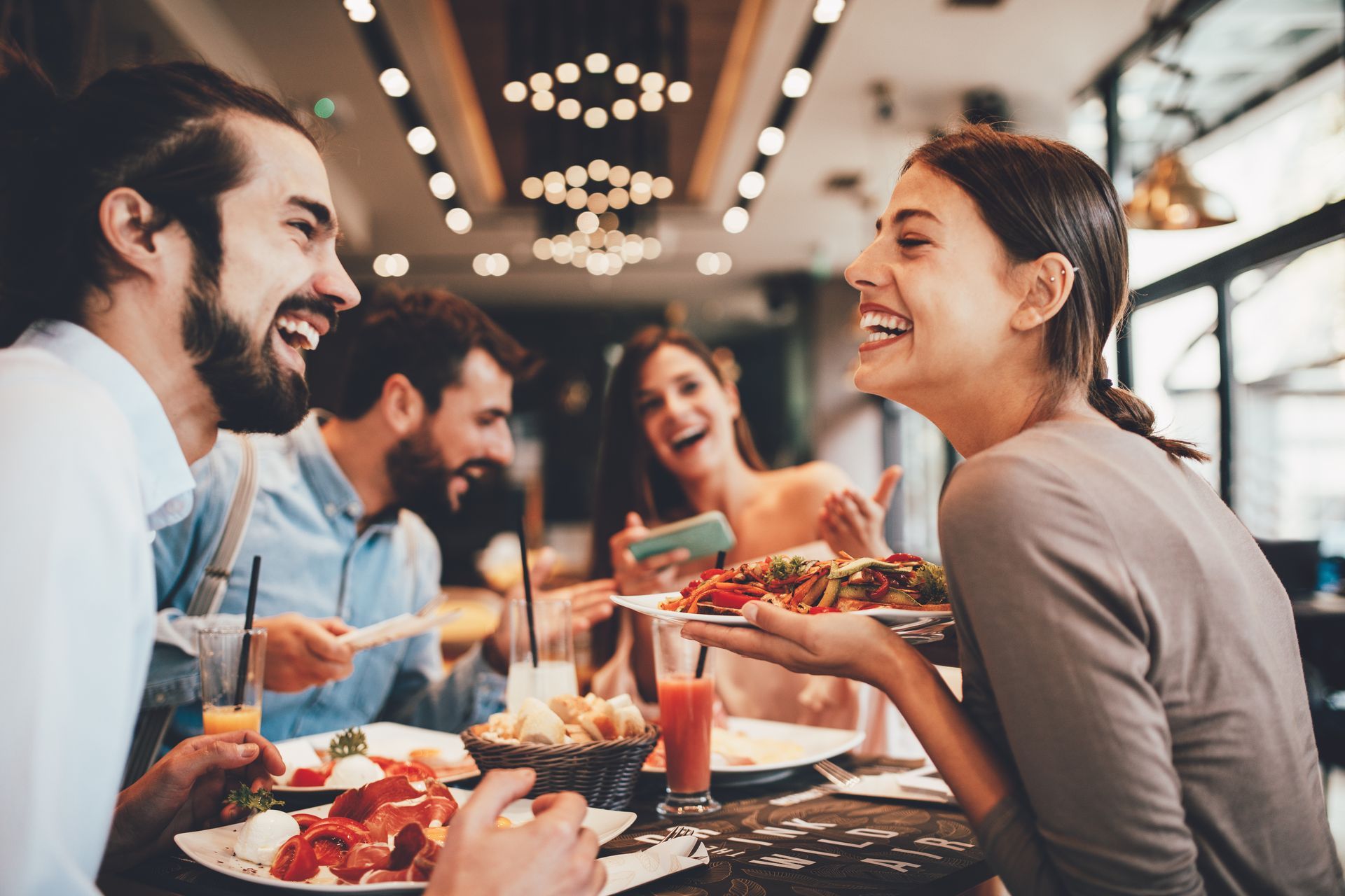 Friends sharing a meal and laughing at a lively restaurant table with candles and pizza
