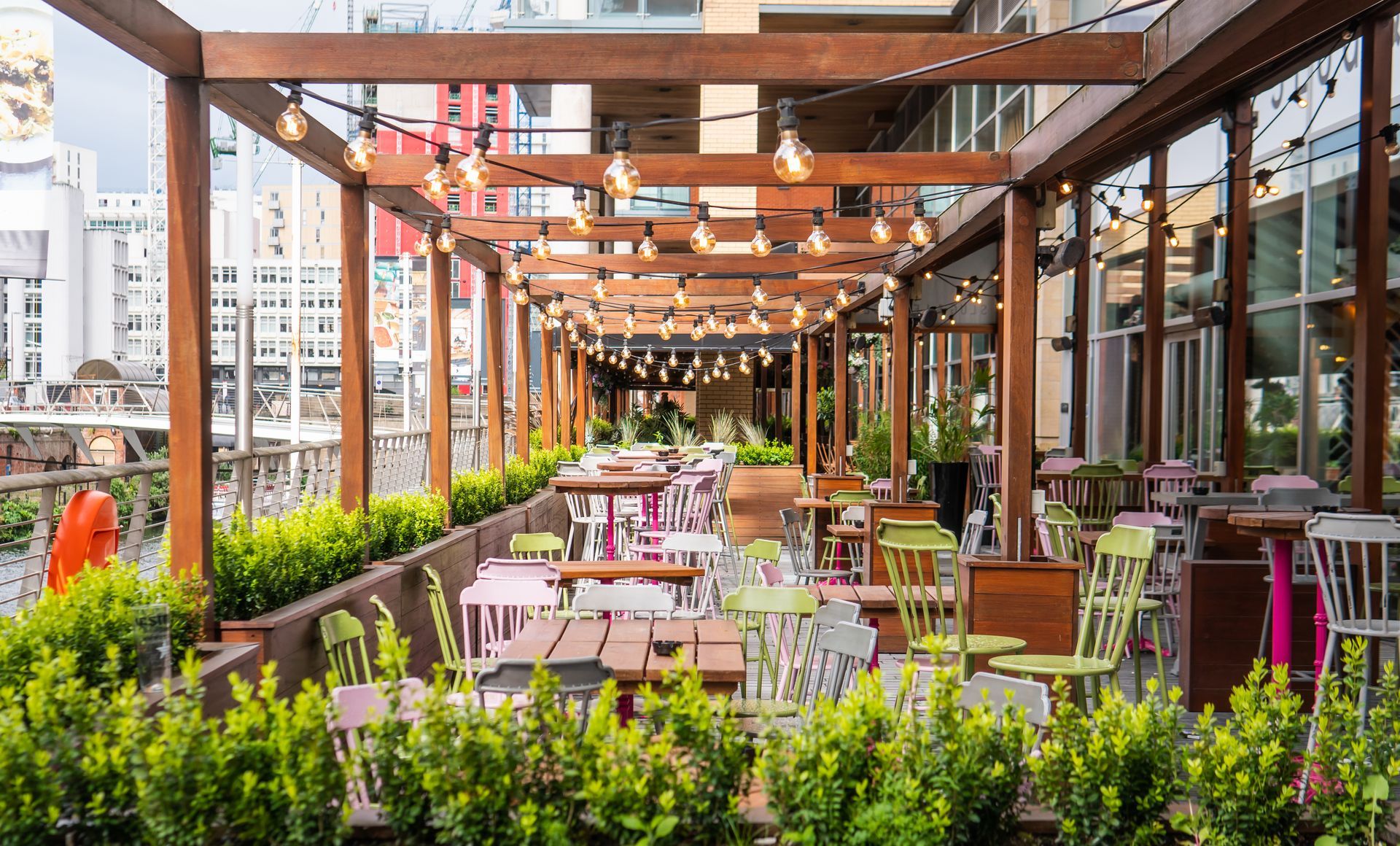 Outdoor rooftop restaurant patio with wooden pergola, pink tables and chairs, and lush green planters