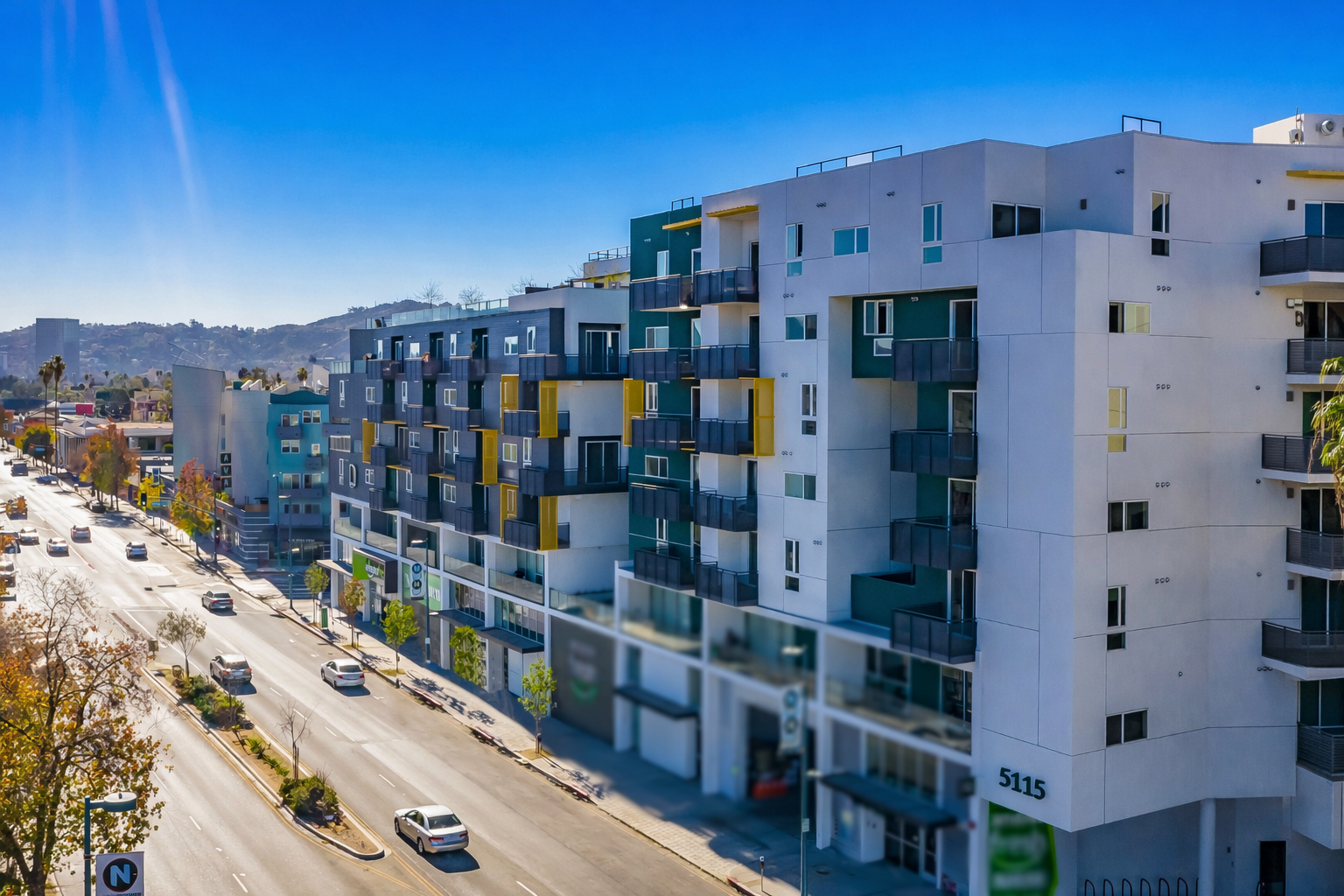 Modern apartment buildings lining a sunlit city street with light traffic and clear blue sky