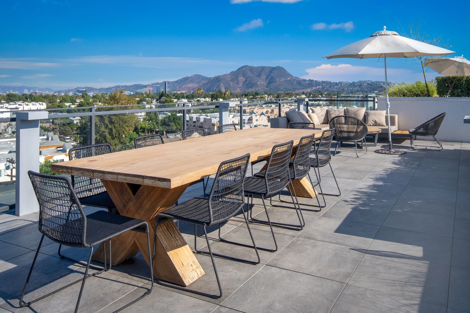 Rooftop patio with a long wooden table, black chairs, umbrella, and city and mountain views