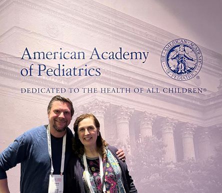 Two people smiling in front of an American Academy of Pediatrics banner at a conference