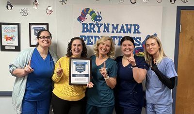 Five people smiling, holding a certificate in front of a Bryn Mawr School wall sign.