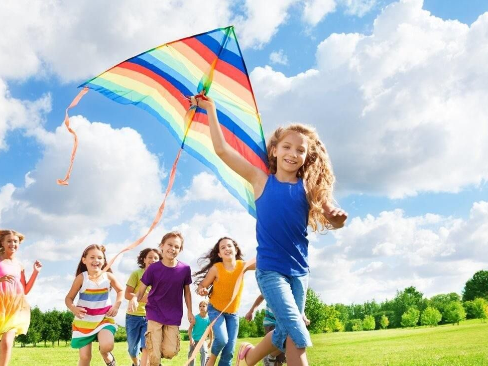 Children running in a grassy field, flying a rainbow kite under a blue sky with clouds