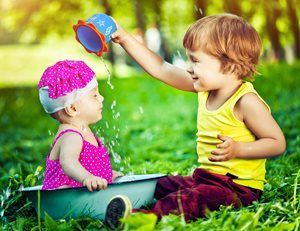 Two toddlers sit in grass, one pouring water from a blue cup onto the other wearing pink and holding a basin.