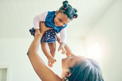 Adult lifting a smiling baby in a blue outfit toward the light indoors