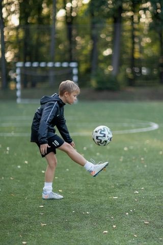 Young soccer player in black kit juggling a ball on a grassy field with trees in the background