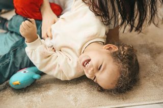 Smiling child lying on a carpet, holding a toy with an adult nearby