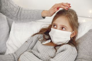 Child wearing a face mask on a couch while an adult checks for fever with a hand on her forehead