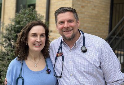 Two people in stethoscopes smiling outdoors near a brick building