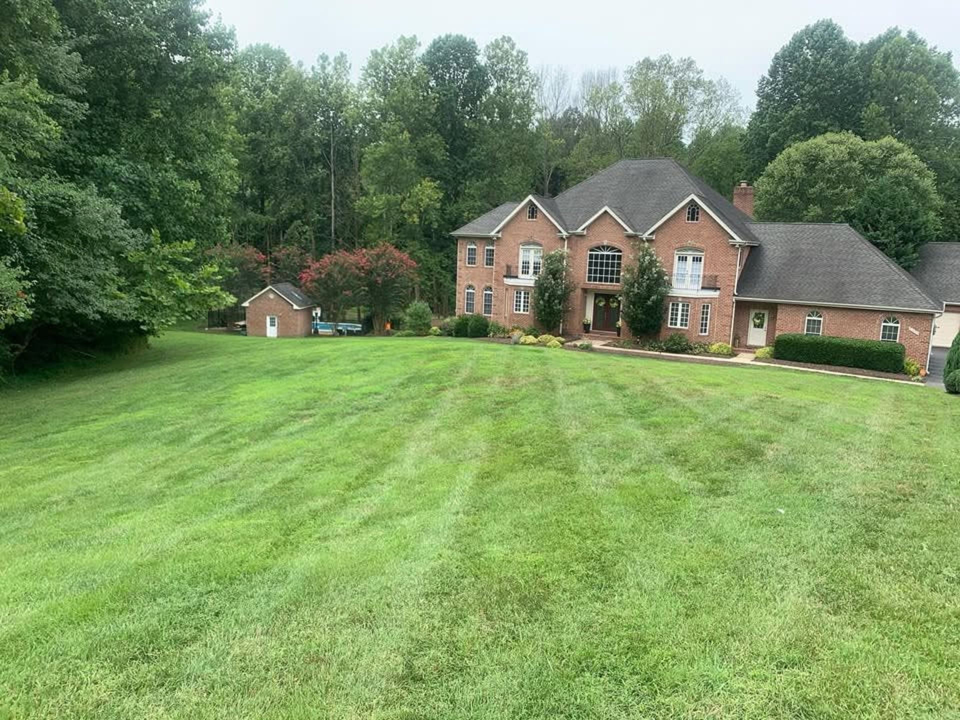 A large, multi-story brick house with a dark roof, surrounded by trees, viewed across a wide, freshly mowed green lawn.