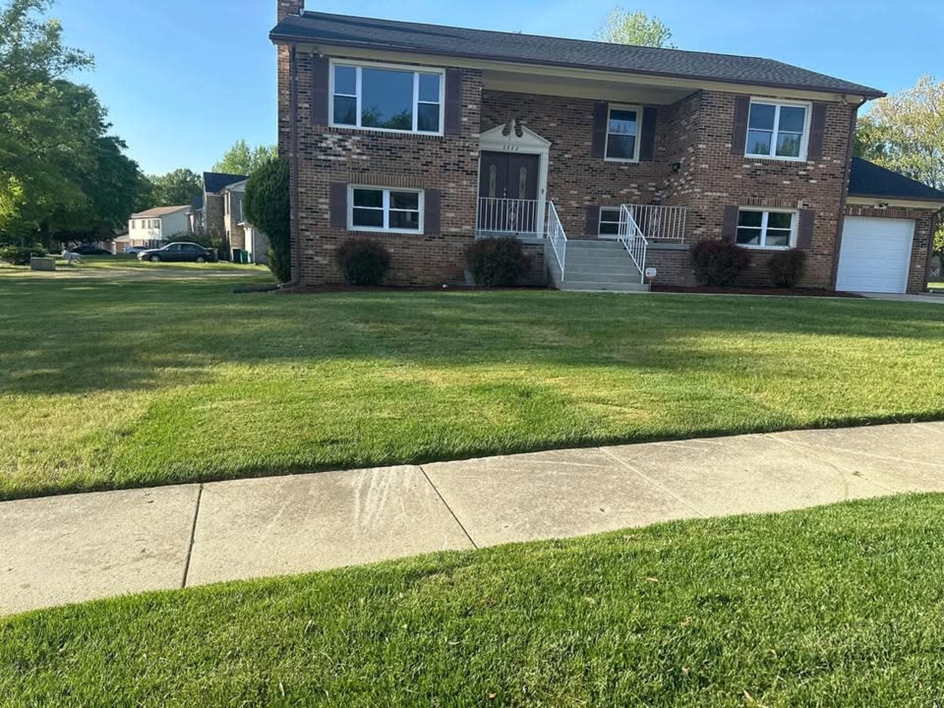 A two-story brick house with a white front door, a set of steps, and a garage, set behind a large green lawn and sidewalk.