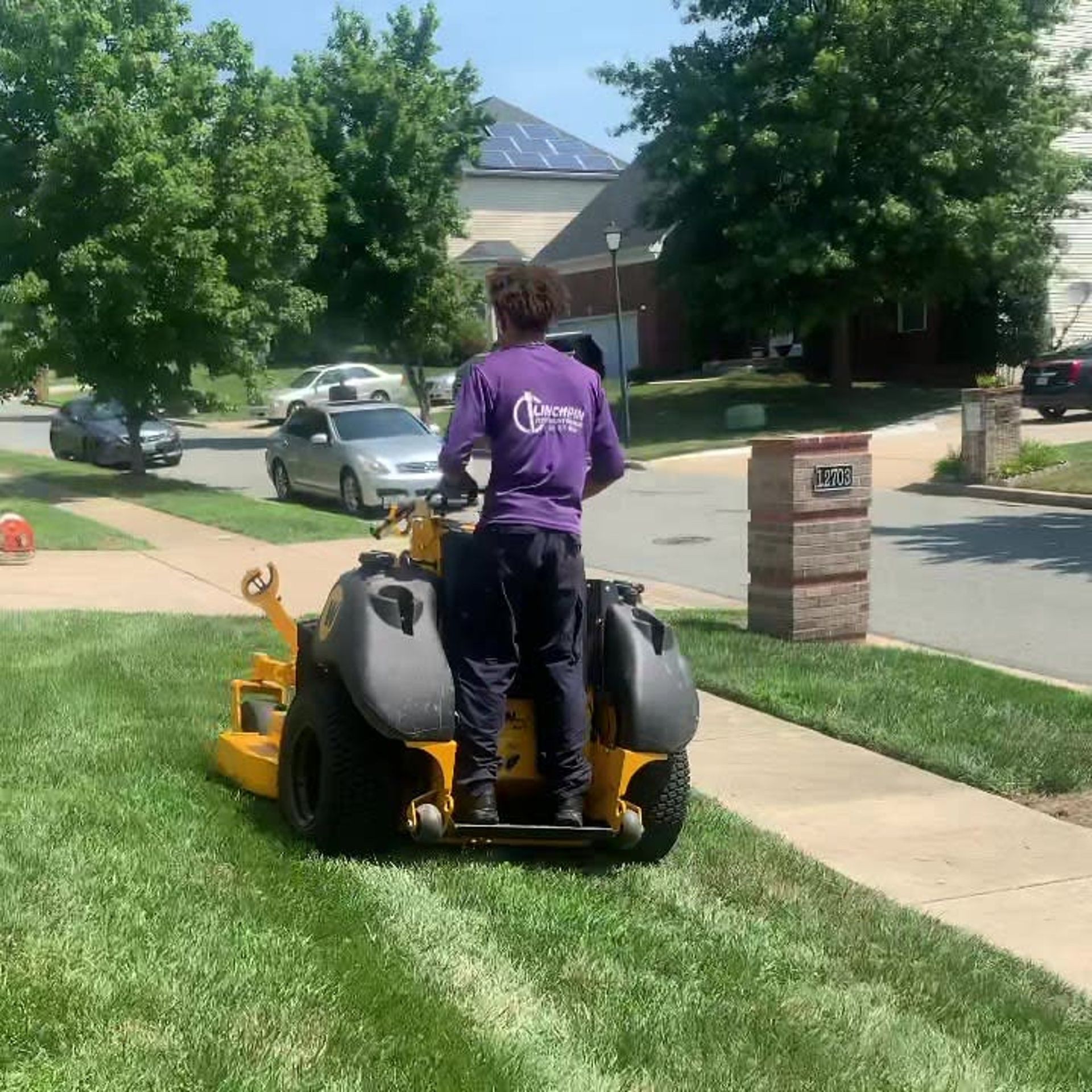 A person in a purple shirt operates a yellow stand-on zero-turn lawn mower on a residential front lawn.