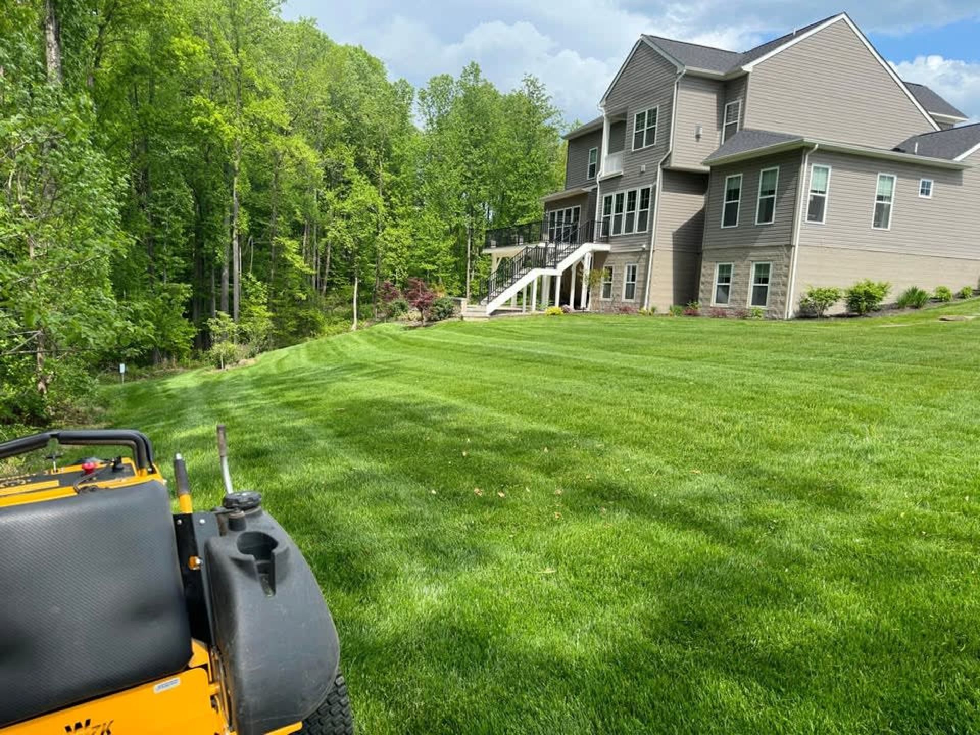 A yellow zero-turn lawn mower in the foreground overlooking a large, freshly mowed green lawn and a suburban house.