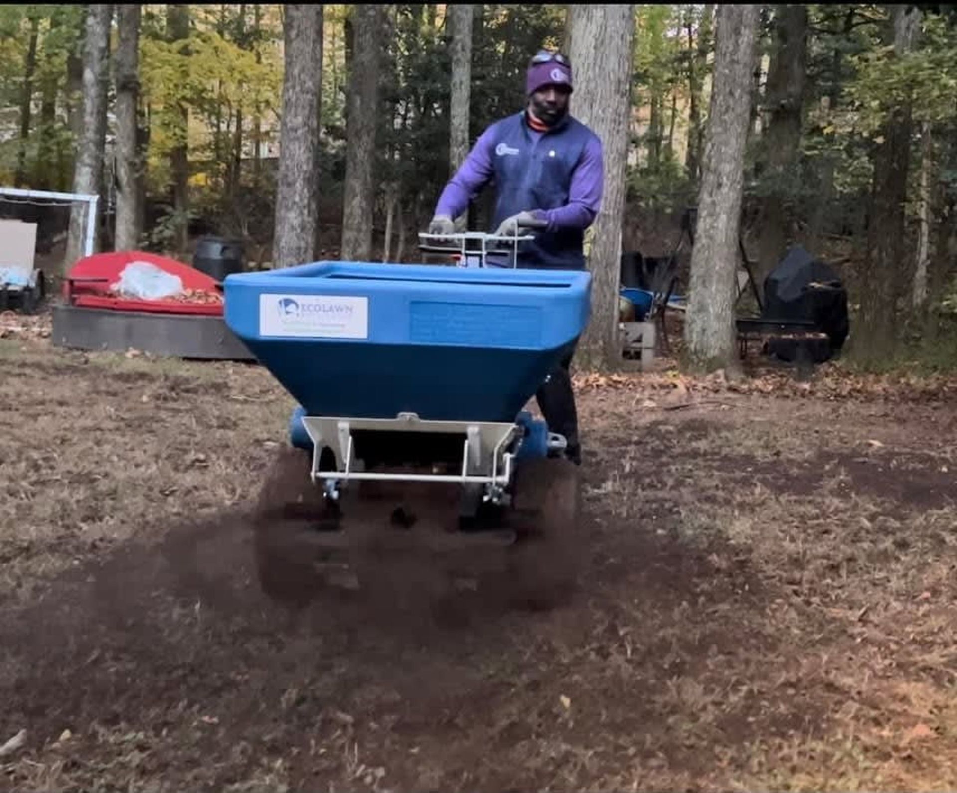 A person uses a blue walk-behind topdresser to spread soil across a lawn in a wooded area.