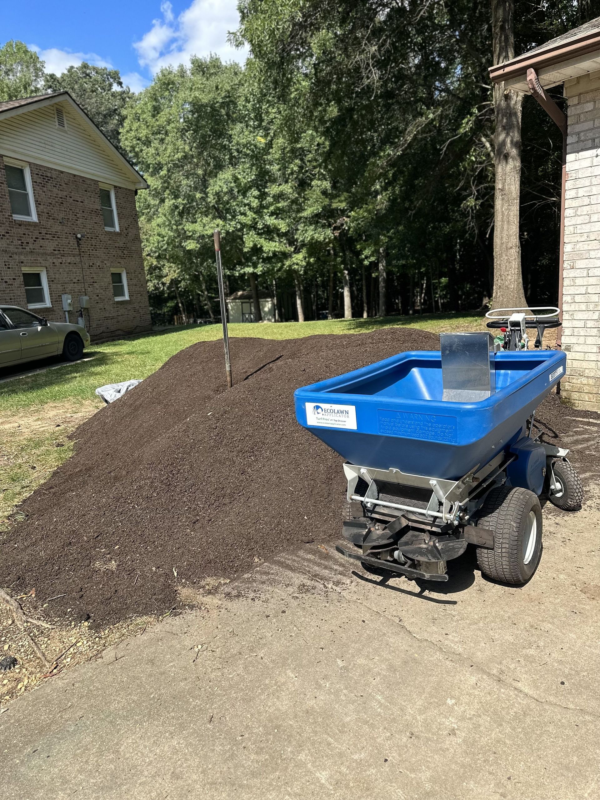 A blue material spreader sits on a driveway next to a large pile of brown mulch with trees and houses in the background.