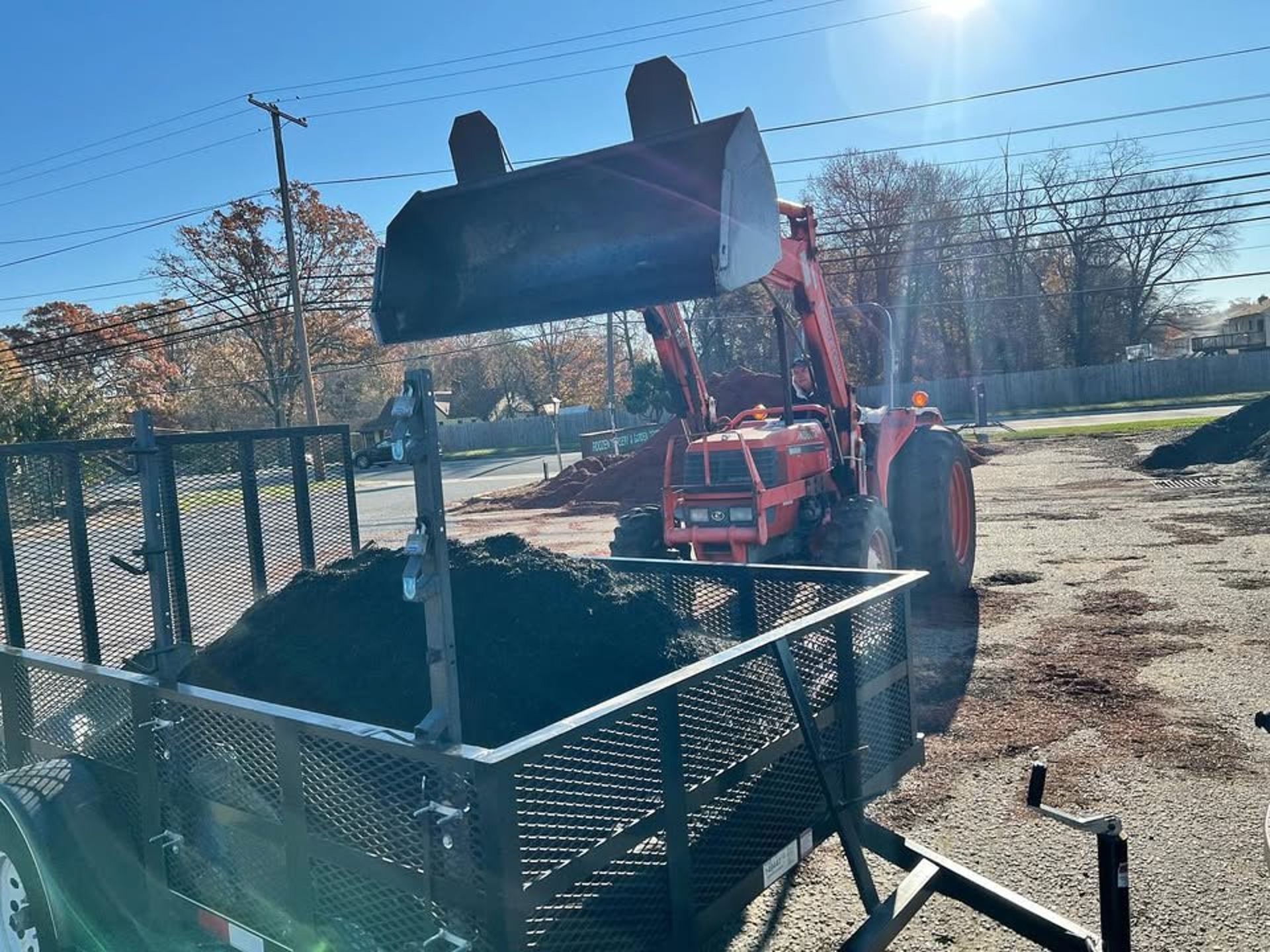 A red tractor with a front loader raised in the air is dumping soil into a utility trailer on a sunny day.