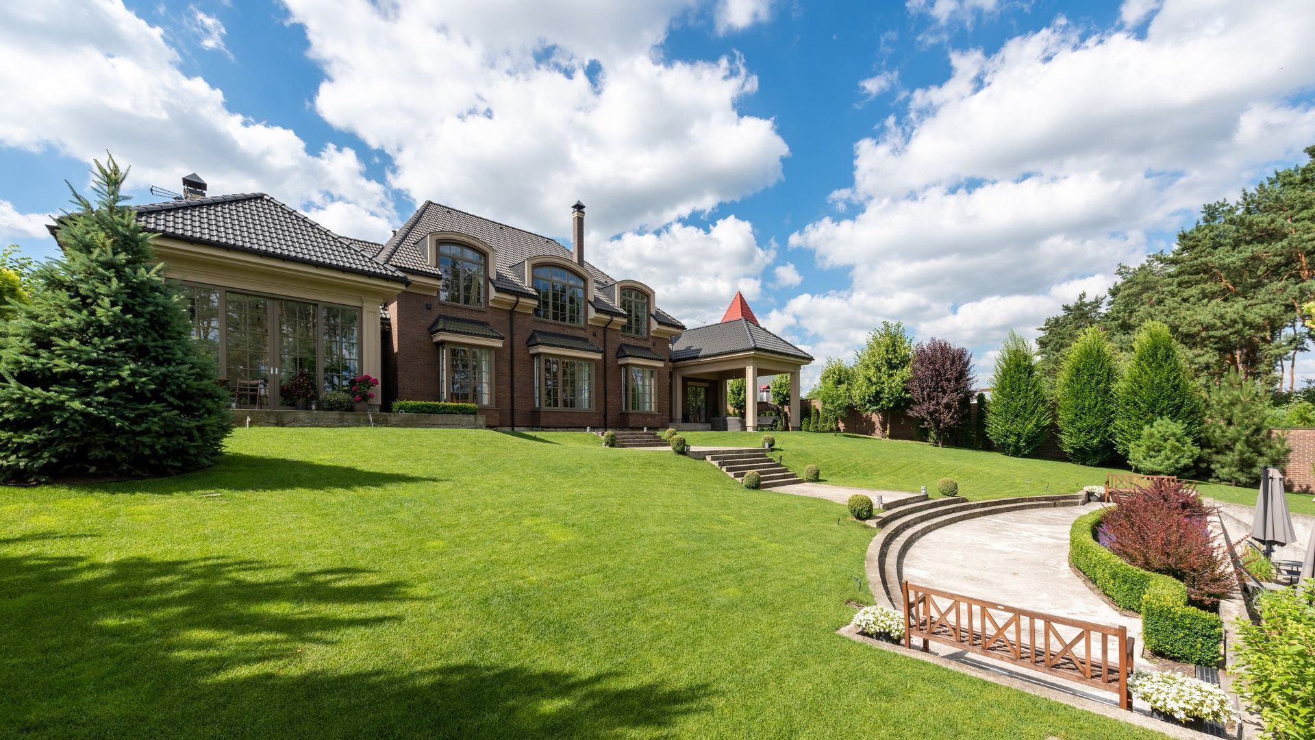 A multi-story brick mansion with a large, manicured lawn, a curved patio, and a stone walkway under a blue, cloudy sky.