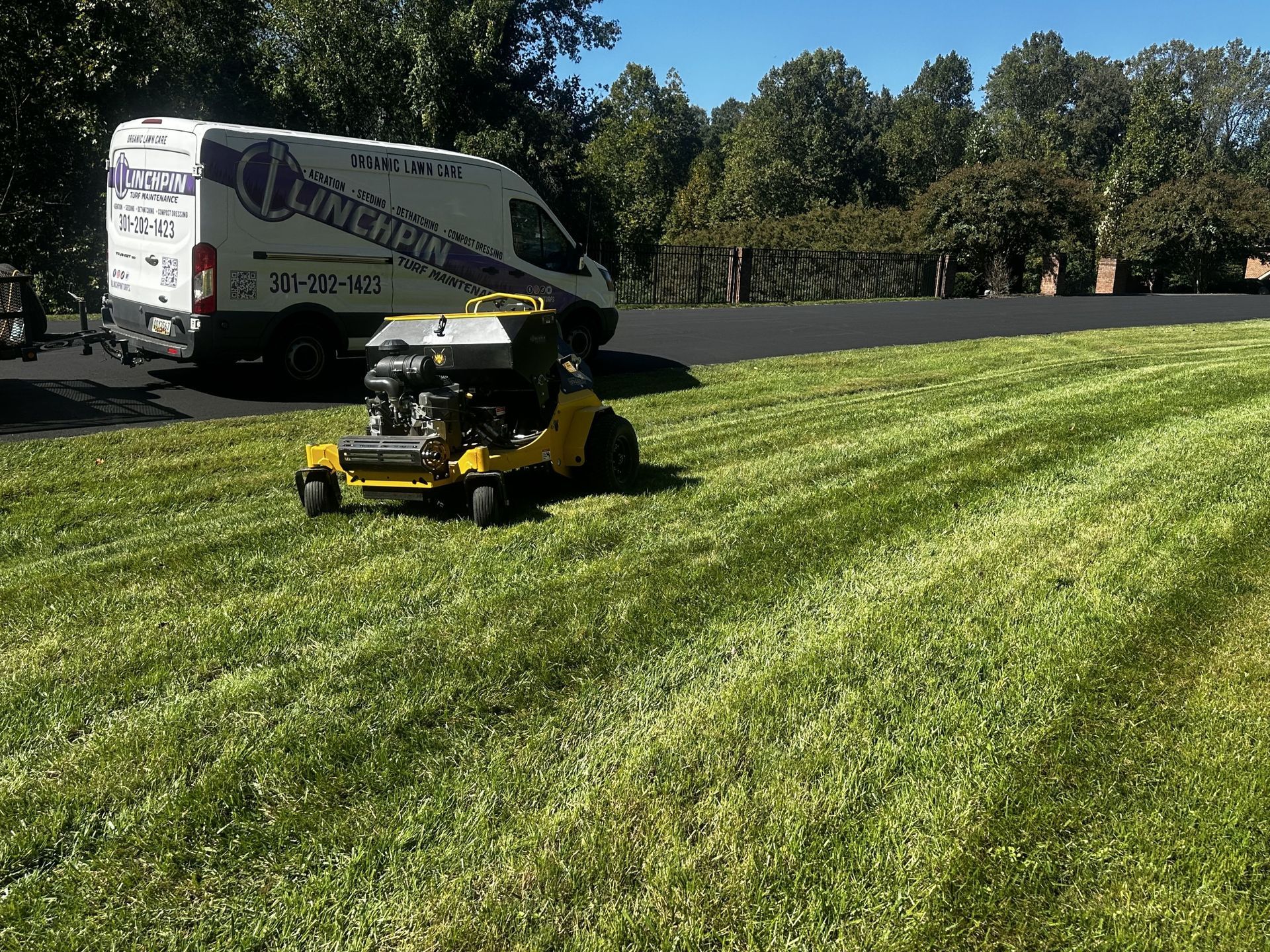 A yellow lawn mower cuts grass on a sunny day next to a parked service van with a business logo.