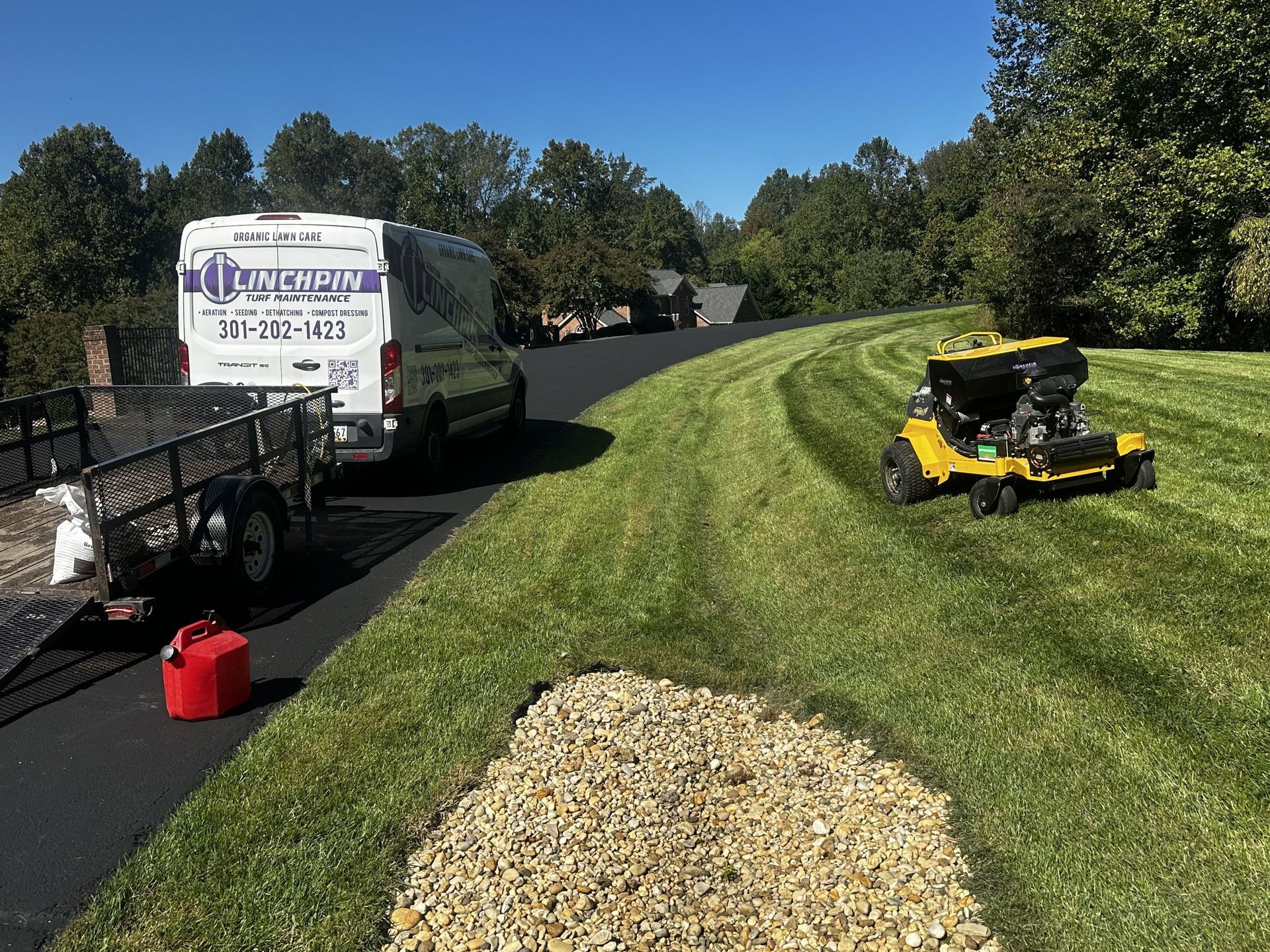 A white work van with a trailer and a yellow riding lawnmower on a grassy verge next to a newly paved asphalt driveway.