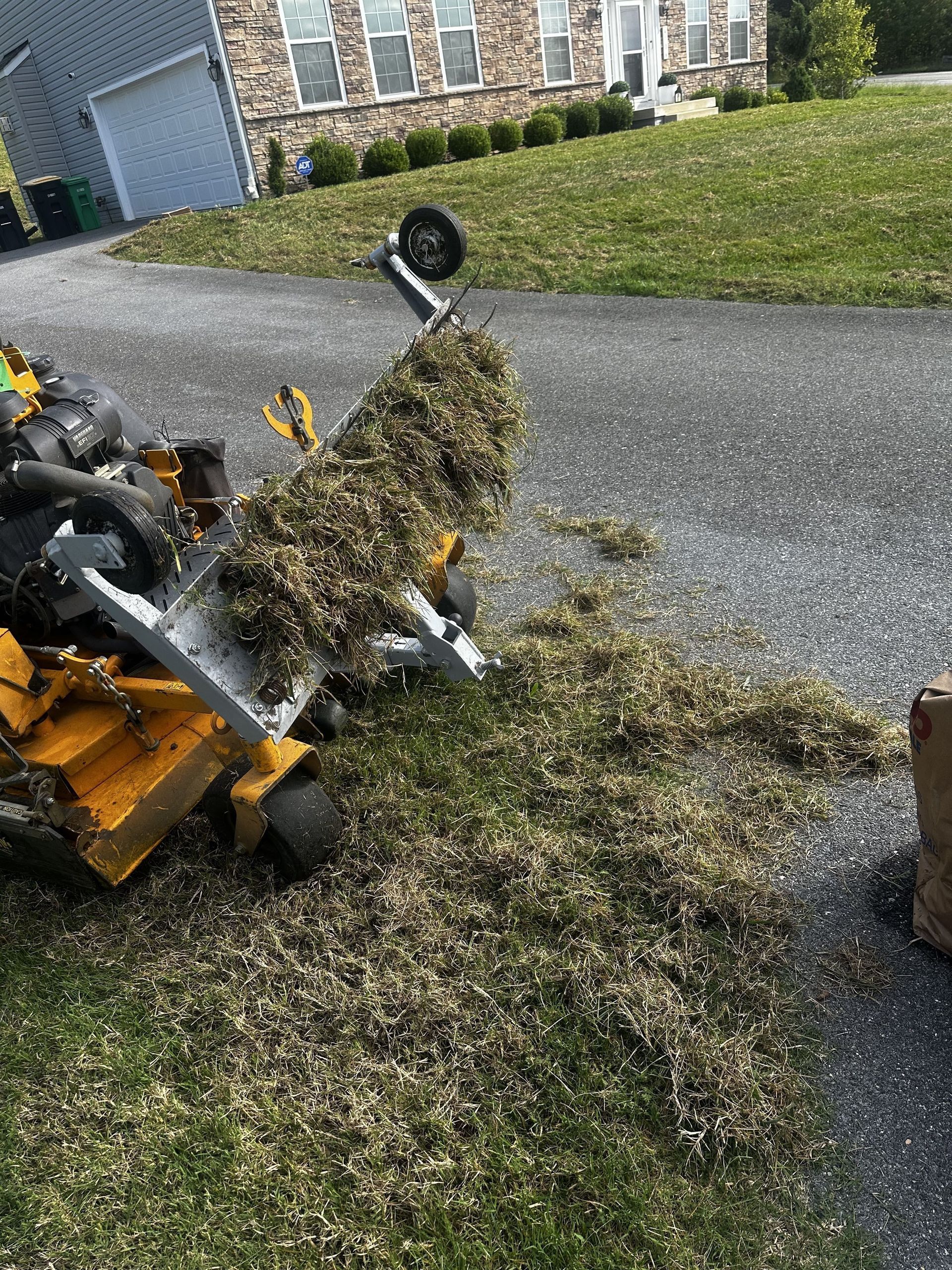 A yellow lawn mower tipped on its side on a driveway, clearing a pile of dead grass and debris.