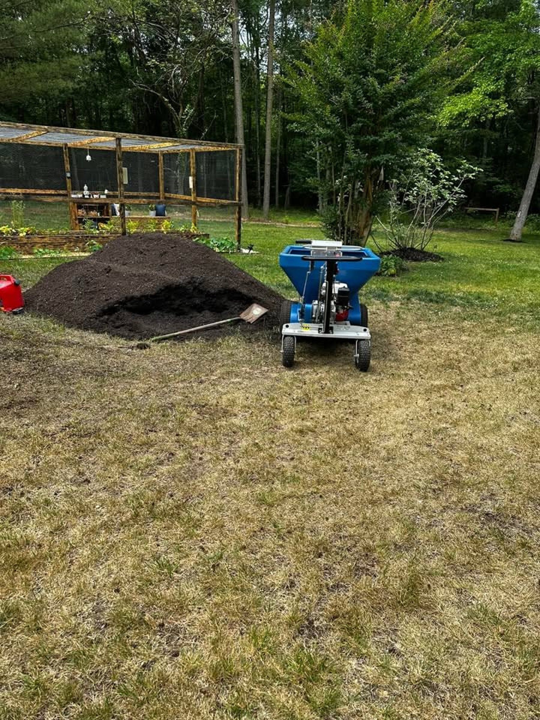 A blue spreader sits on a grassy lawn next to a large pile of dark mulch, with a chicken coop in the background.