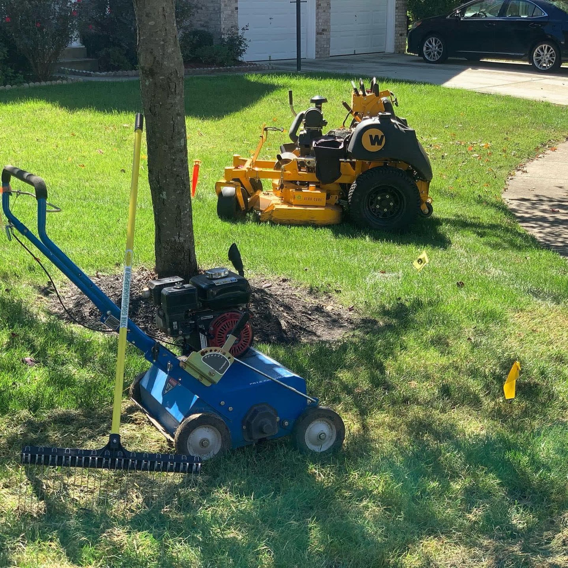 A blue lawn dethatcher and a yellow zero-turn mower sit on a green lawn near a tree on a sunny day.