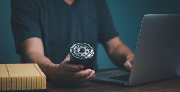A man is holding an automotive oil filter and buying it online from a laptop computer. 