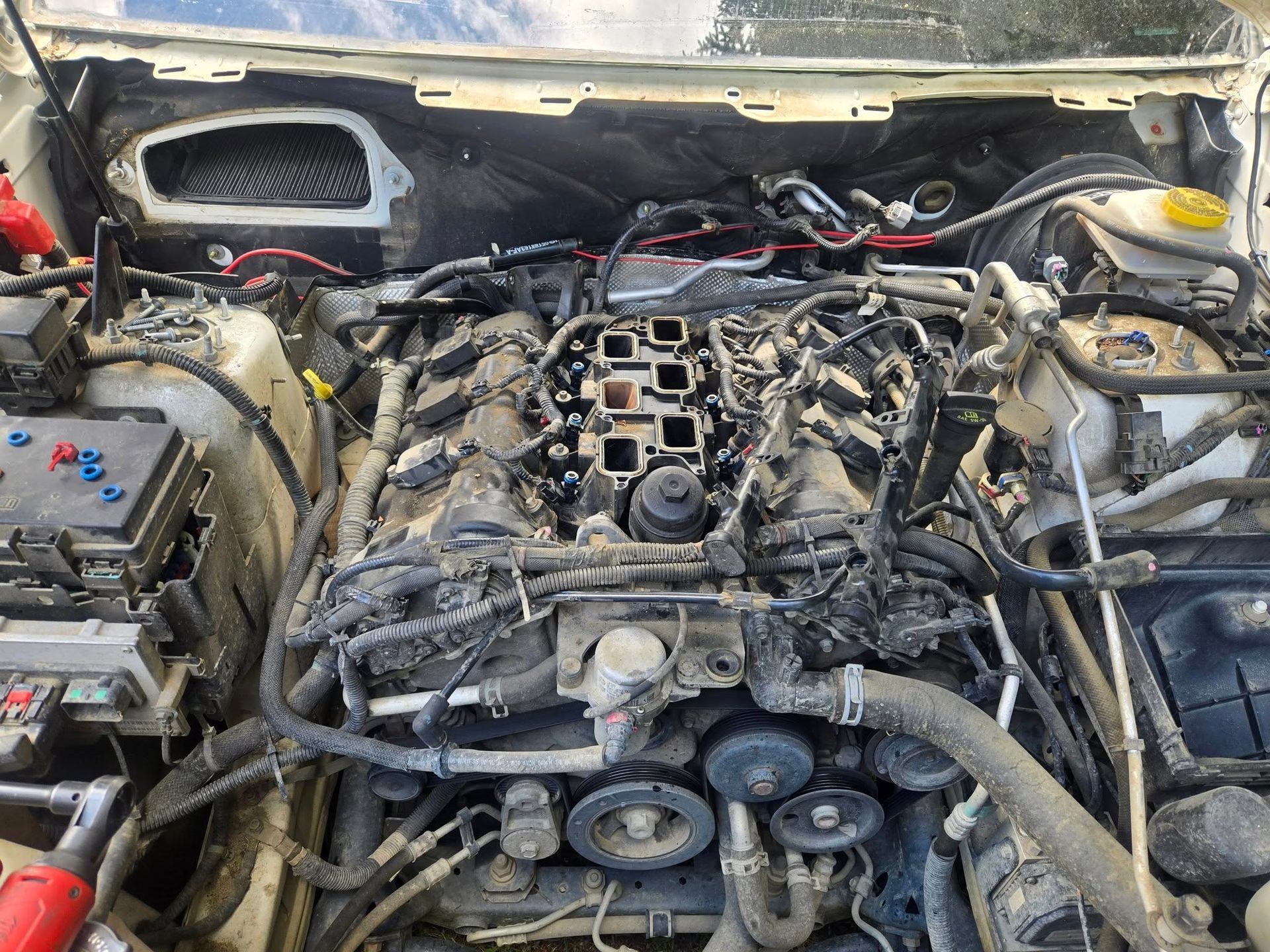 Open car engine bay with exposed engine components, likely undergoing repair. Wires, tubes, and other parts are visible.