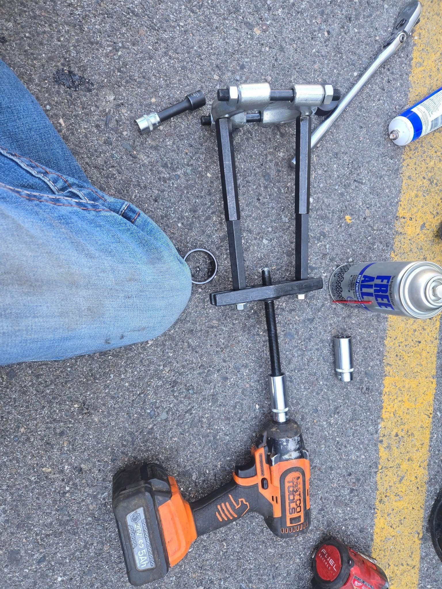 An orange and black power drill attached to a suspension tool, with jeans and asphalt visible.