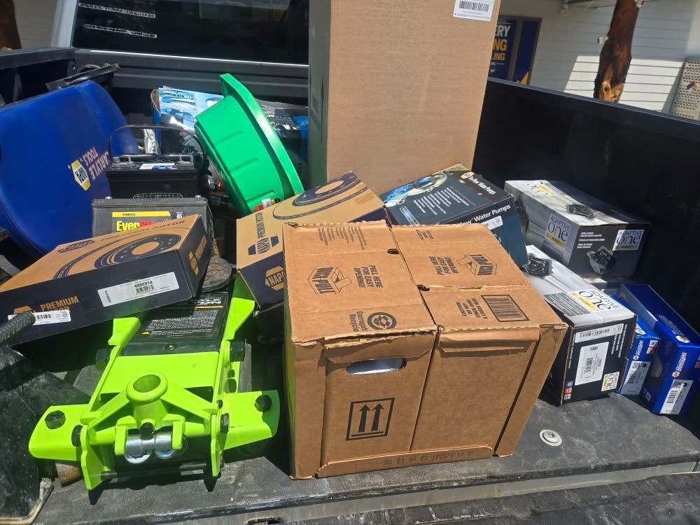 Truck bed filled with car parts, boxes, a green jack, and other equipment.