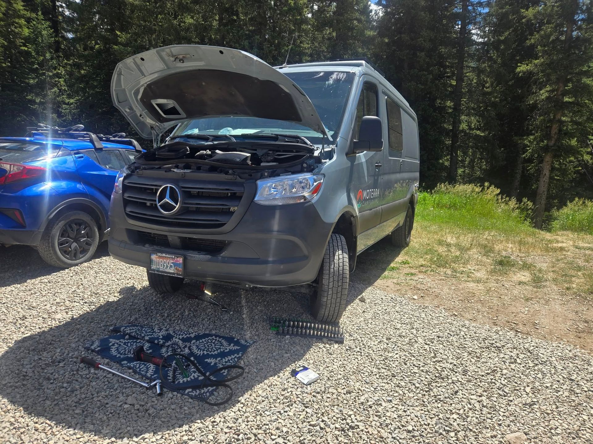 A gray van with its hood open sits in a gravel parking area, tools scattered on the ground. A blue car is parked beside it.