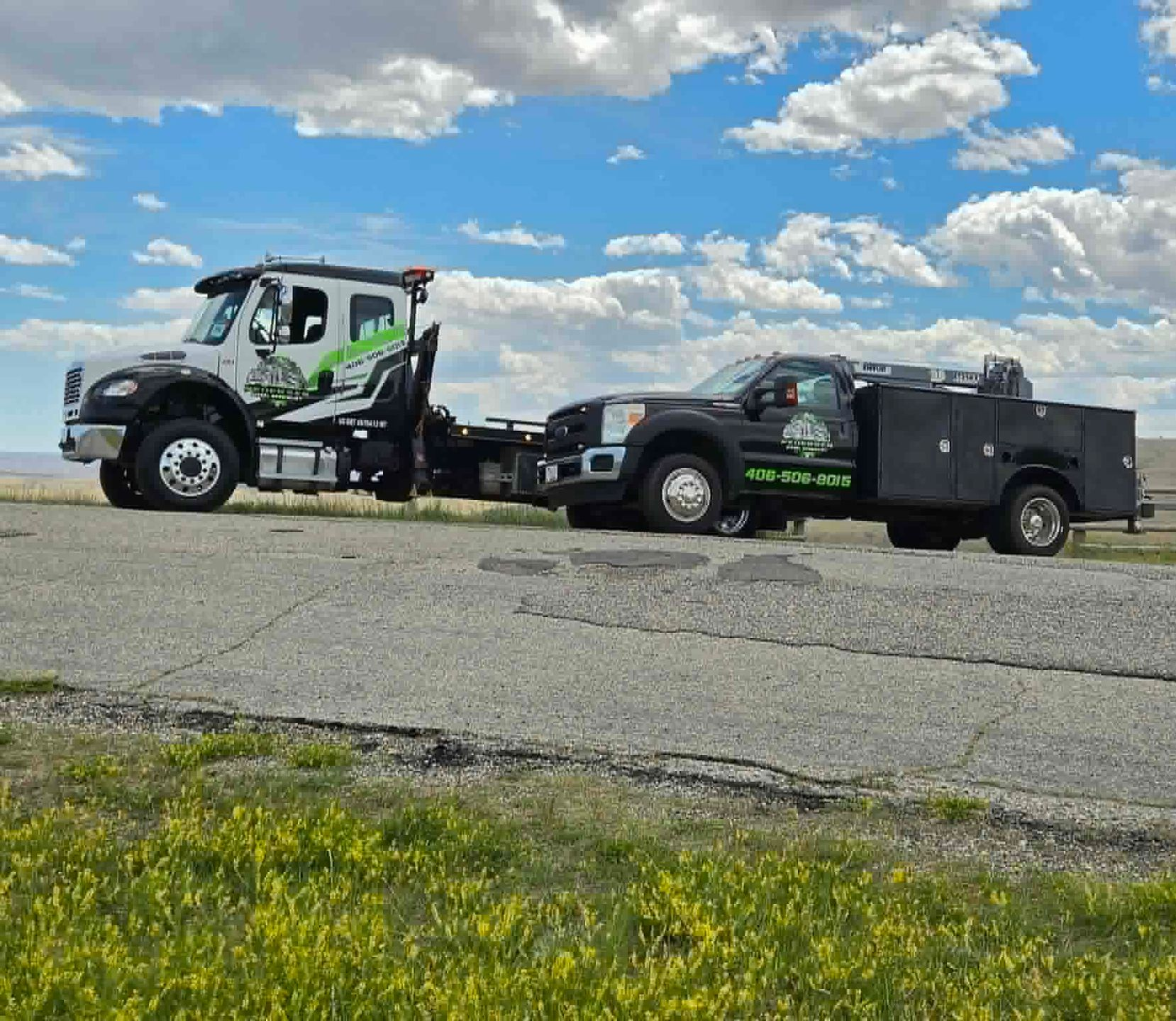 Two tow trucks parked on a cracked lot under a blue sky with scattered clouds
